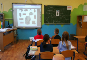 Zdjęcie przedstawia fragment sali lekcyjnej i siedzących w ławkach uczniów. Dzieci oglądają wyświetlane na tablicy multimedialnej ilustracje elementów wyposażenia i uzbrojenia Powstańców Warszawskich. Fotografia wykonana jest z perspektywy tylnego widoku sali.