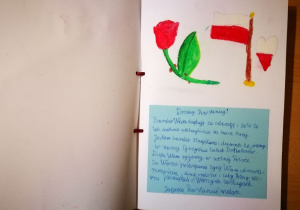 Zdjęcie przedstawia kartkę w albumu dla Powstańców Warszawskich wykonaną przez ucznia. W górnej części kartki namalowany jest czerwony kwiat, a tuż obok flaga Polski i biało-czerwone serce. W dolnej części kartki wklejone są życzenia dla Powstańców, które napisane zostały na niebieskim kartonie.