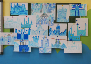 Zdjęcie przedstawia wisząca na ścianie w sali lekcyjnej zieloną tablicę kredową. Na tablicy znajdują się prace plastyczne dzieci przedstawiające zamki i pałace ,,Królowej Śniegu". W pracach dominuje kolor niebieski i szary.
