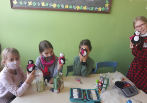 Na zdjęciu znajdują się 4 uczennice klasy 2b. W rękach trzymają wykonane na warsztatach pingwiny. Pingwiny mają wokół szyi różowe szaliki. Za dziewczynkami widać zieloną ścianę oraz kawałek tablicy.
