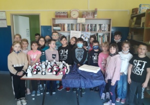Uczniowie klasy 2b stojący w bibliotece szkolnej, przed uczniami znajduje się stół, na którym stoją wykonane przez uczniów pingwiny. Za uczniami znajdują się regały z książkami.