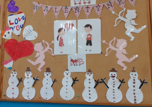 Zdjęcie przedstawia prace dzieci: kolorowe serca, trzy Amorki z łukiem w dłoni, 7 bałwanków. W górnej części tablicy widoczna jest girlanda z napisem "Walentynki" oraz obrazki przedstawiające dzieci.