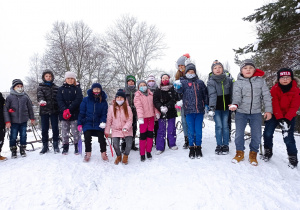 Zdjęcie przedstawia grupę uczniów z klas 3, chłopców i dziewczynki, ubranych w zimowe kurtki, czapki i rękawiczki, stojących na górce w pobliżu szkoły. Uczniowie trzymają w dłoniach śnieżne kulki, w tle na zdjęciu są widoczne drzewa oraz budynki mieszkalne. Niektóre z dzieci mają sanki i jabłuszka do zjeżdżania z górki.